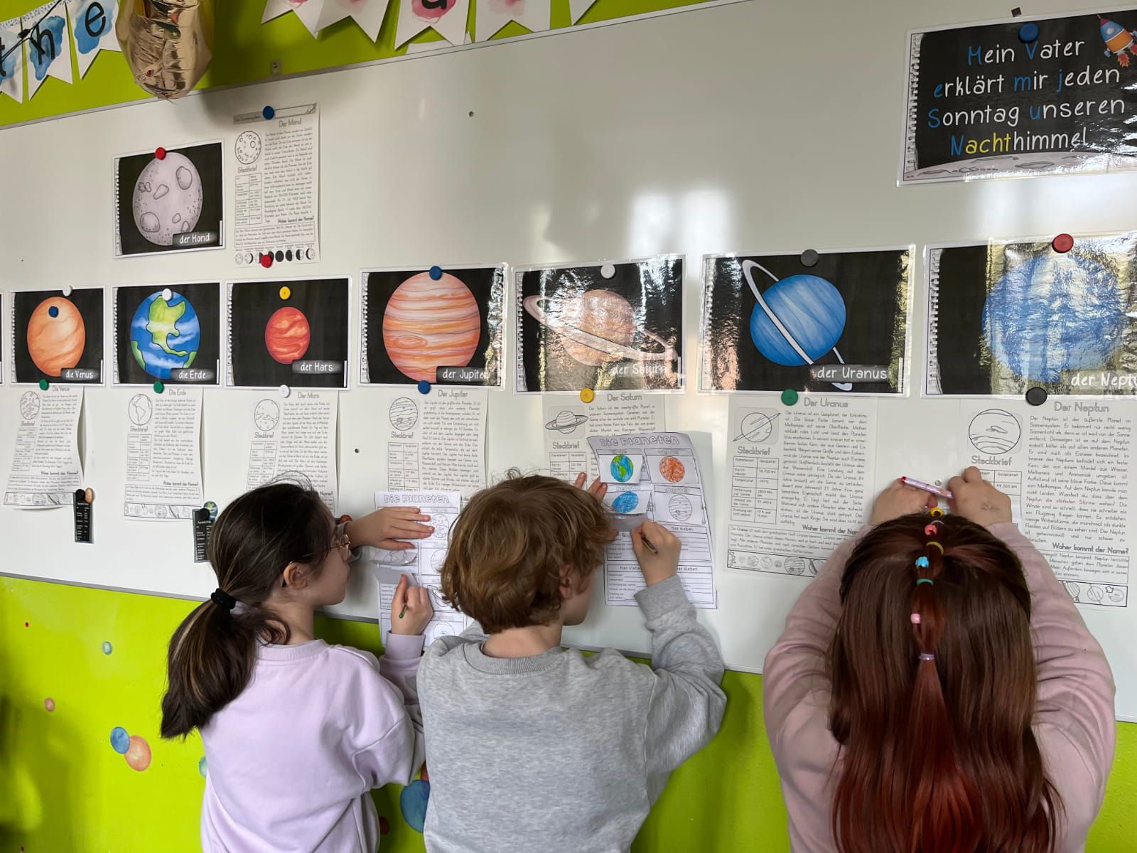 drei Kinder schreiben in Arbeitsblätter an einer Magnettafel